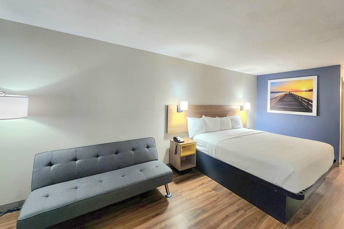 A hotel room with a bed, a sofa, a nightstand with a lamp, and a painting on the wall.