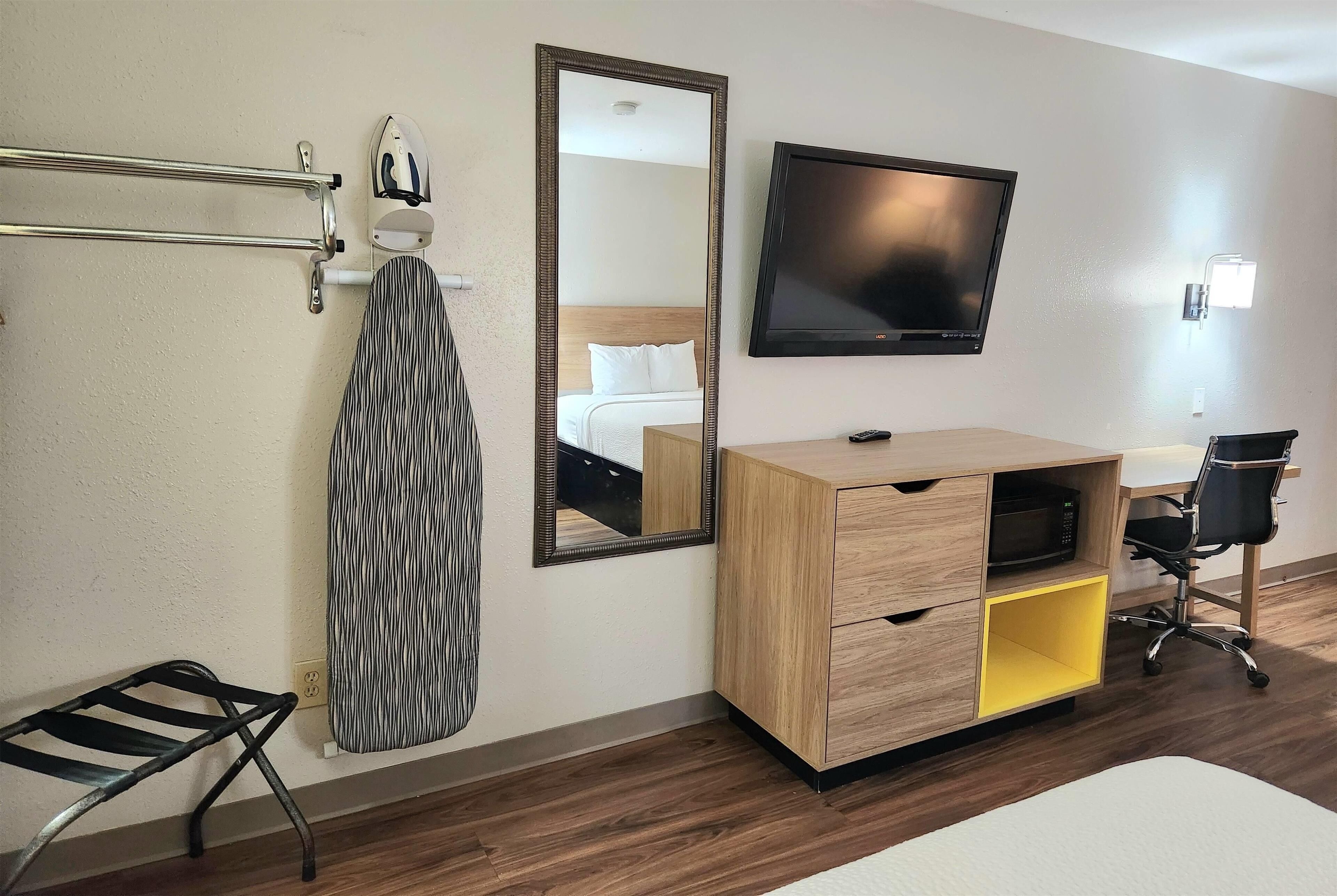 A hotel room with a wooden desk, a flat-screen TV, a mirror, a coat rack, and a chair.