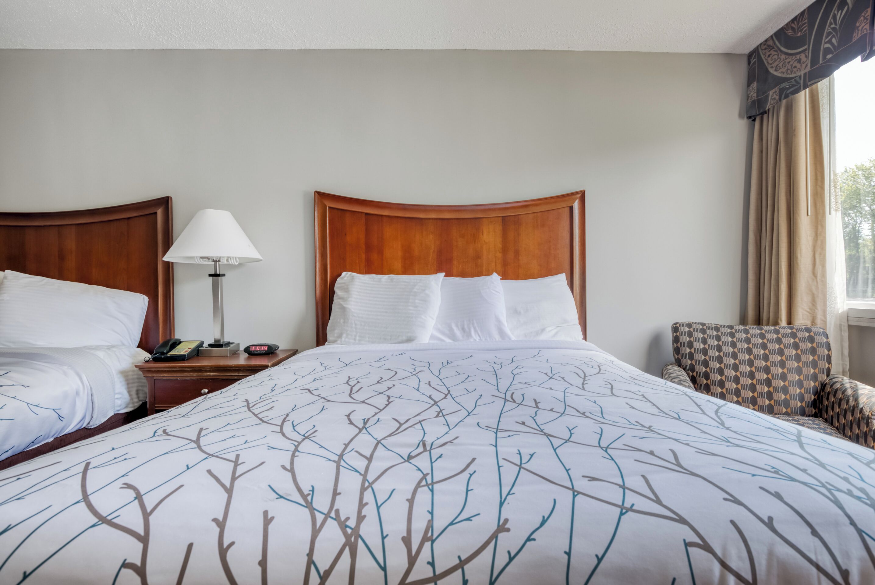 A hotel room with two beds, a bedside table with a lamp, a chair, and a window with curtains.