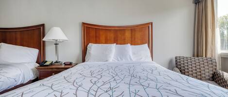 A hotel room with two beds, a bedside table with a lamp, a chair, and a window with curtains.