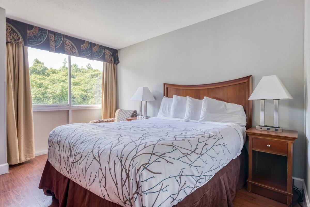 A hotel room with a large bed, two bedside tables with lamps, a nightstand, and a window with curtains.