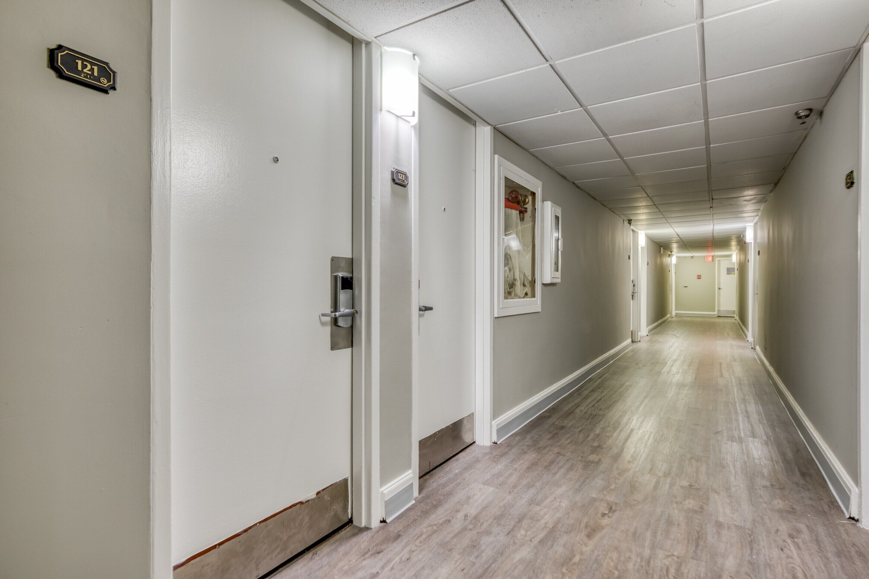 A long, narrow hallway with white walls and wooden flooring. There are doors on the left, one of which is open. A sign above one door reads "121".