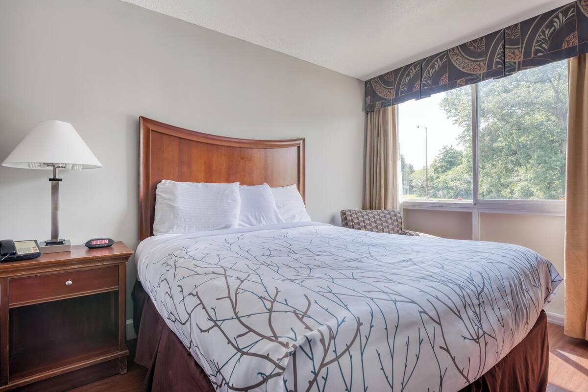 A hotel room with a wooden headboard, a bed with a white and brown patterned bedspread, a bedside lamp, a telephone, and a window with curtains.