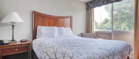 A hotel room with a wooden headboard, a bed with a white and brown patterned bedspread, a bedside lamp, a telephone, and a window with curtains.