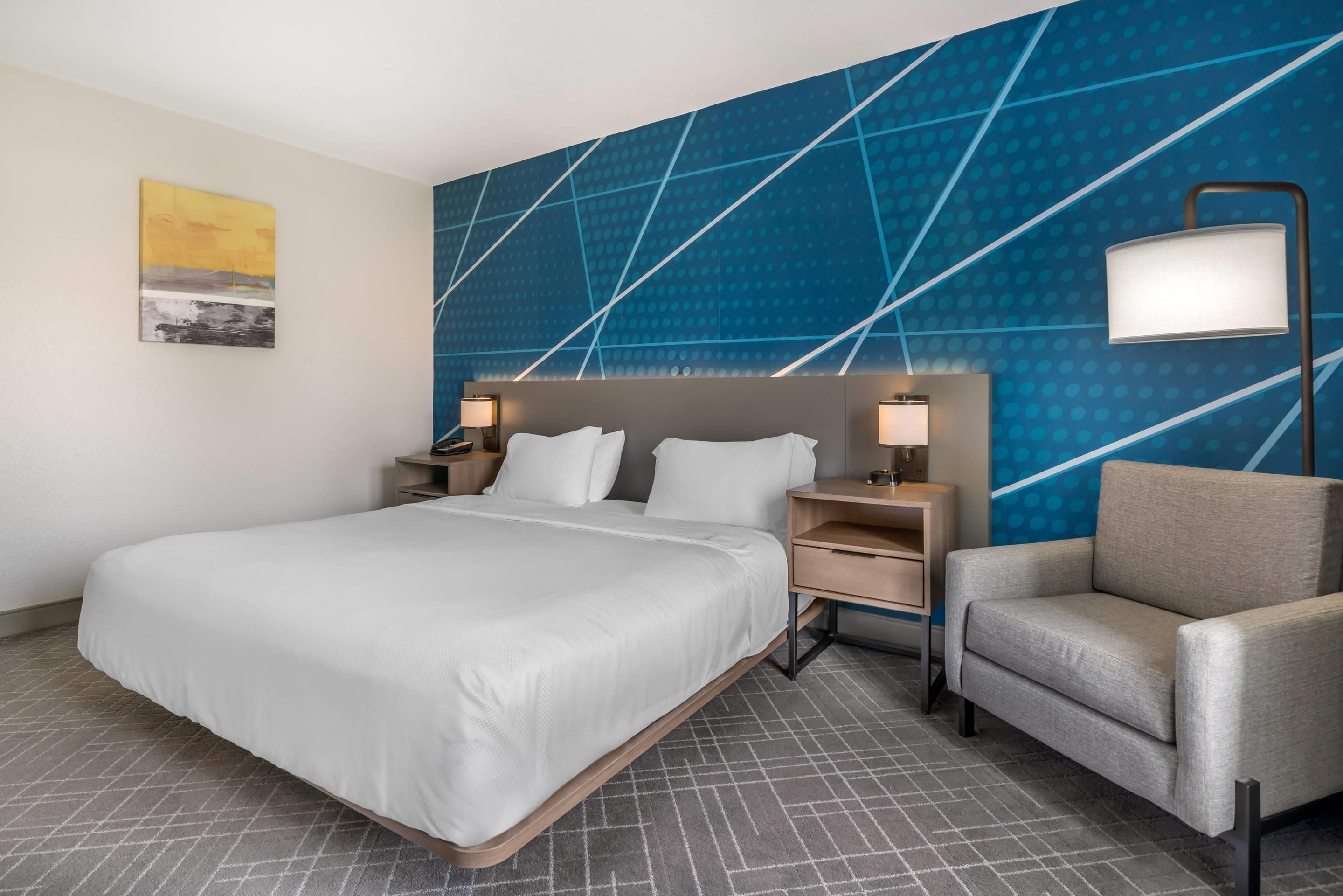 A hotel room with a large bed, a desk, a chair, and a wall with a geometric pattern.