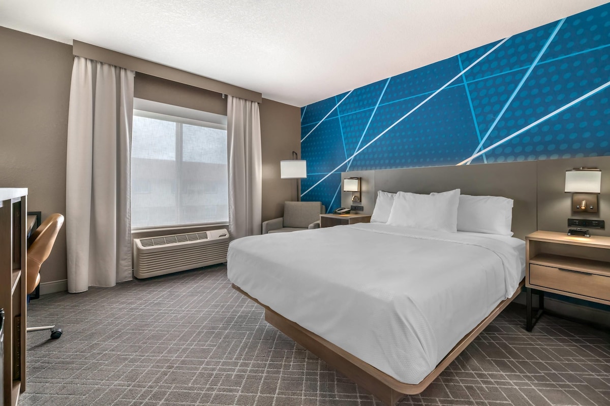 A hotel room with a large bed, a desk, a chair, a window with curtains, and a wall with a geometric pattern.
