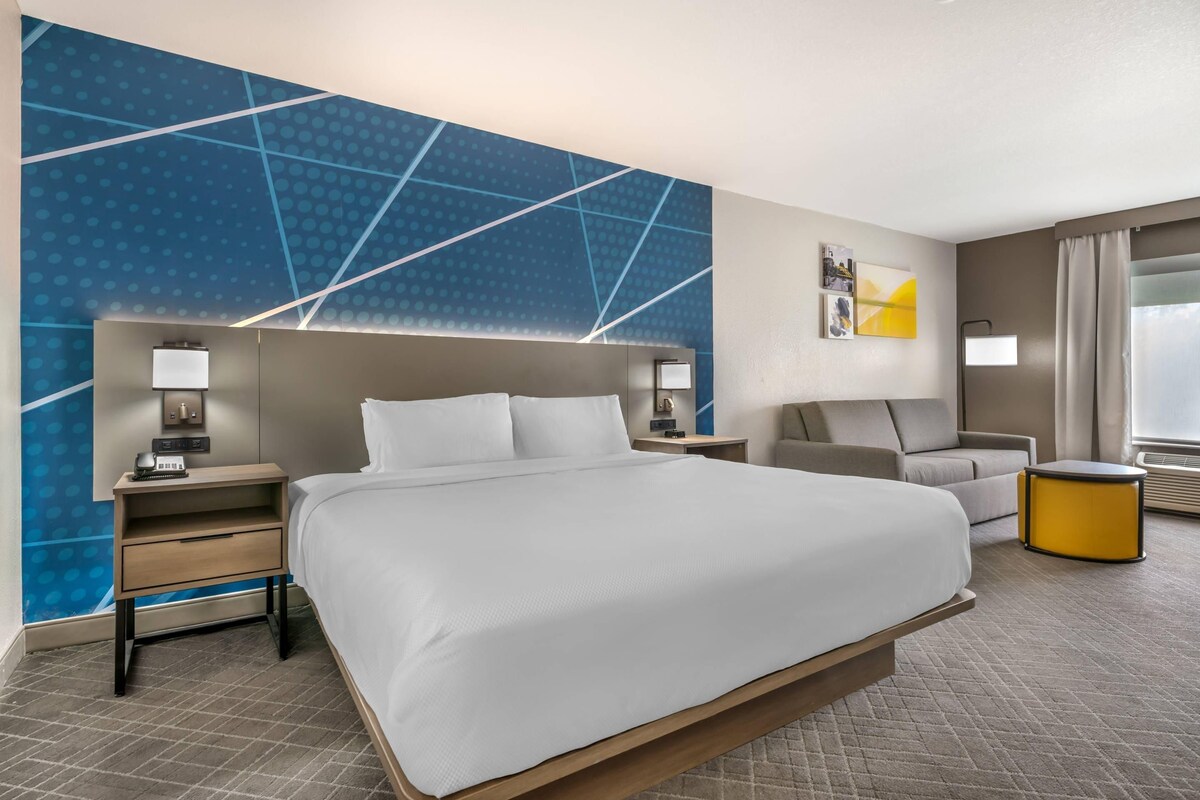 A hotel room with a large bed, a desk, a chair, a small round table, and a wall with a geometric pattern.