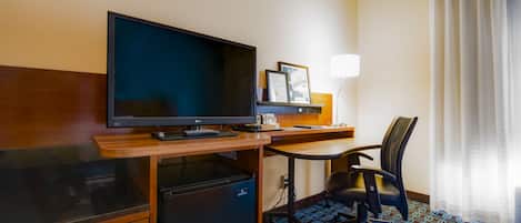 A hotel room with a wooden desk, a television, a refrigerator, a chair, and a patterned carpet.