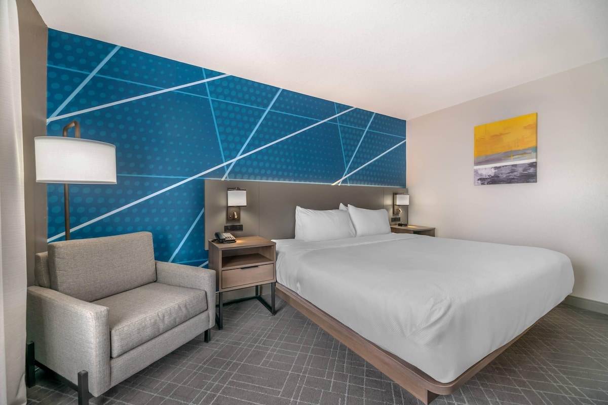 A hotel room with a large bed, a chair, a nightstand with a lamp, and a wall with a geometric pattern.