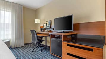 A hotel room with a bed, a desk with a computer, a television, a small refrigerator, and a chair.
