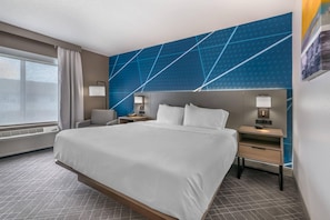 A hotel room with a large bed, two bedside tables, a chair, a window with curtains, and a blue geometric wall design.