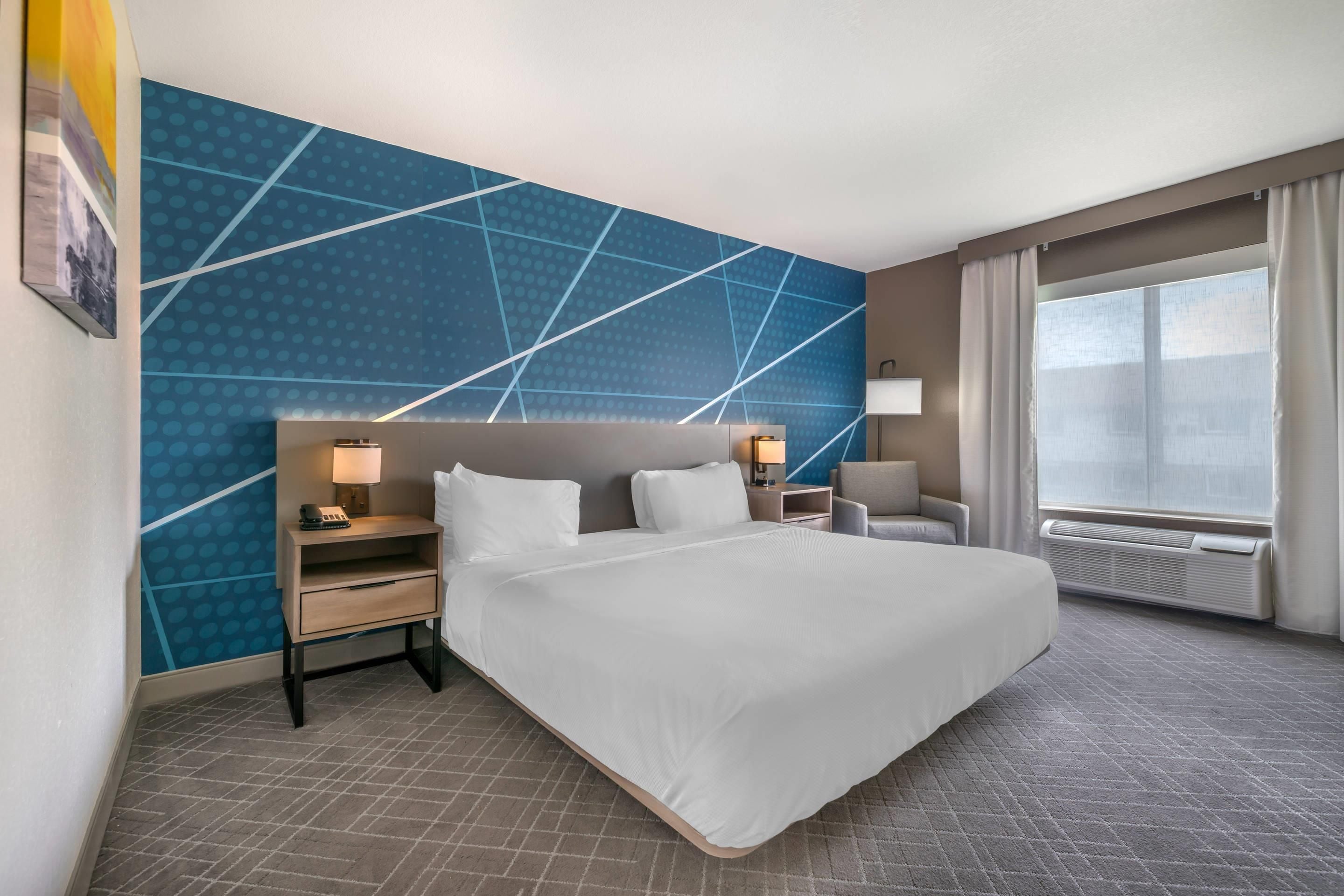 A hotel room with a large bed, two bedside tables with lamps, a chair, a large window with curtains, and a blue patterned wall.