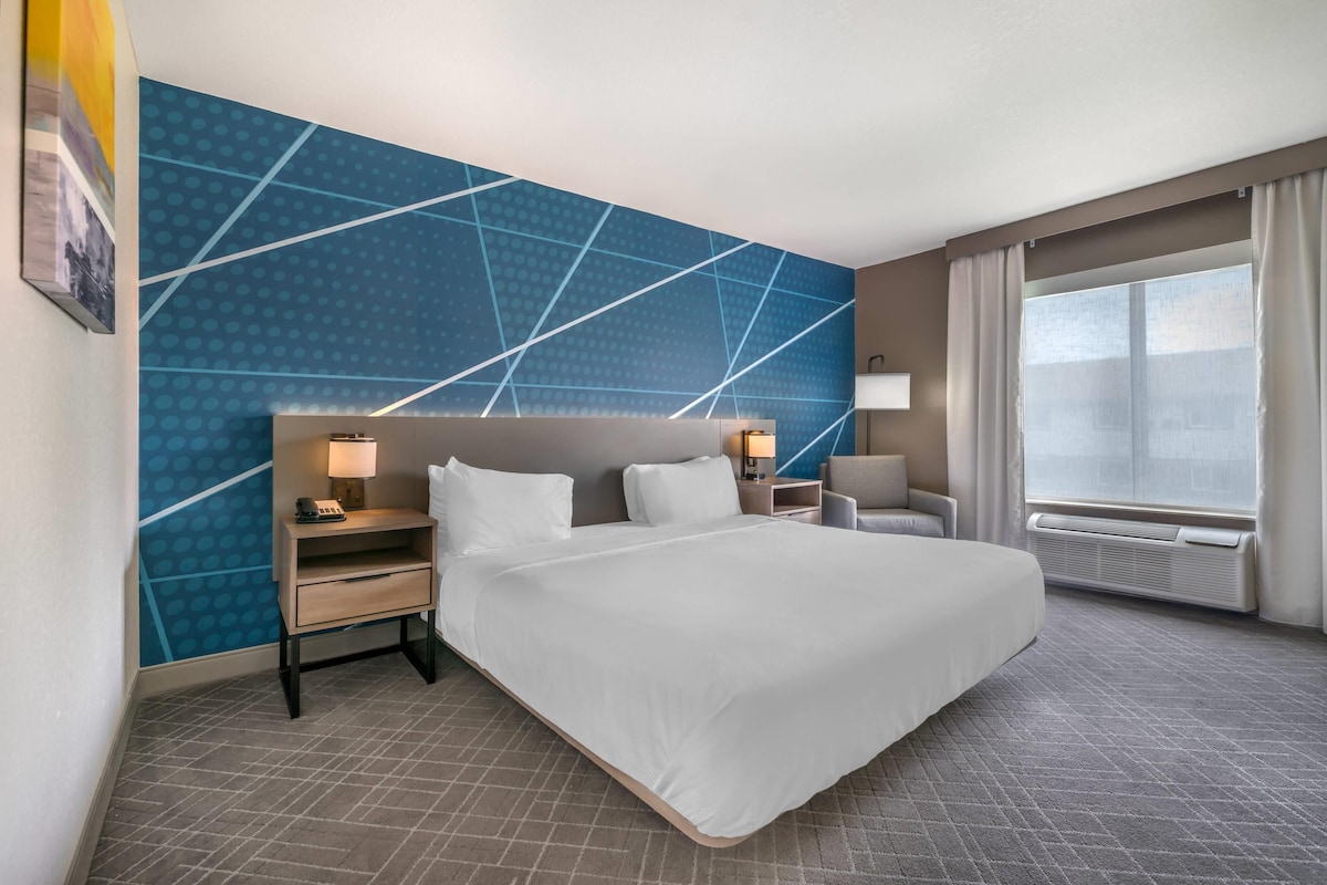 A hotel room with a large bed, two bedside tables with lamps, a chair, a large window with curtains, and a blue patterned wall.