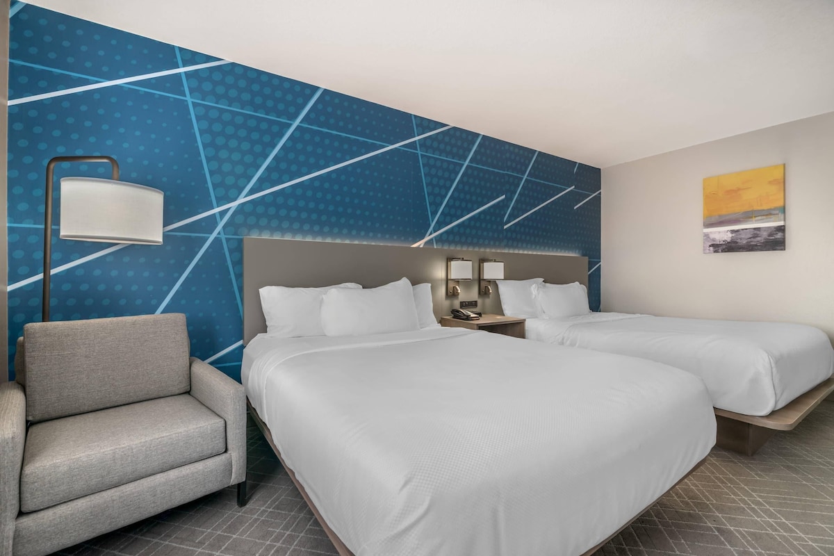 A hotel room with two beds, a chair, a nightstand with a lamp, and a wall with a geometric pattern.