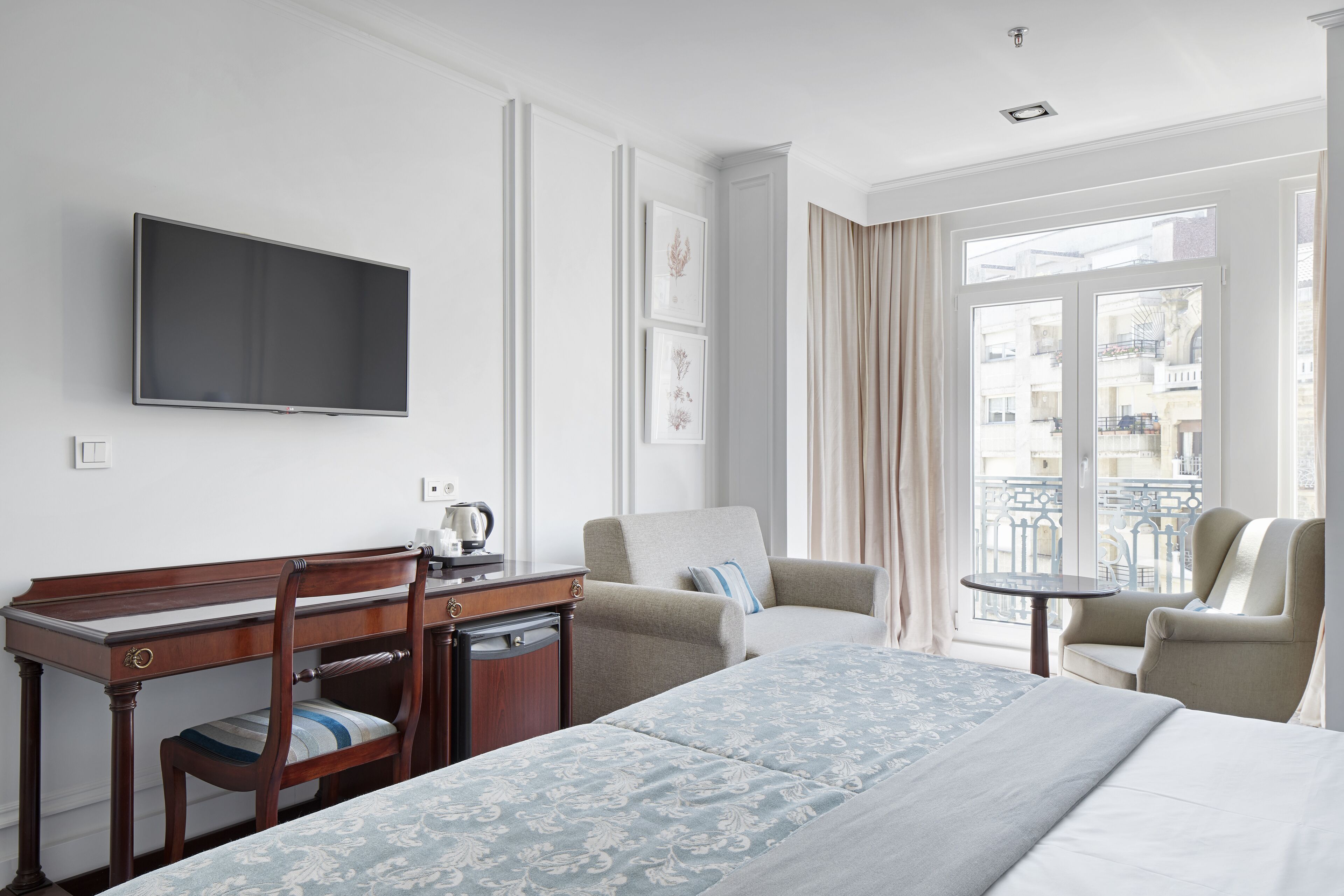 A hotel room with a bed, a desk, a chair, a television, and a balcony with curtains.
