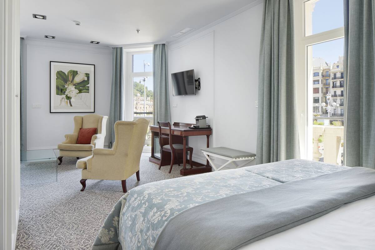 Een hotelkamer met een bed, twee fauteuils, een bureau, een televisie en een groot raam met gordijnen.