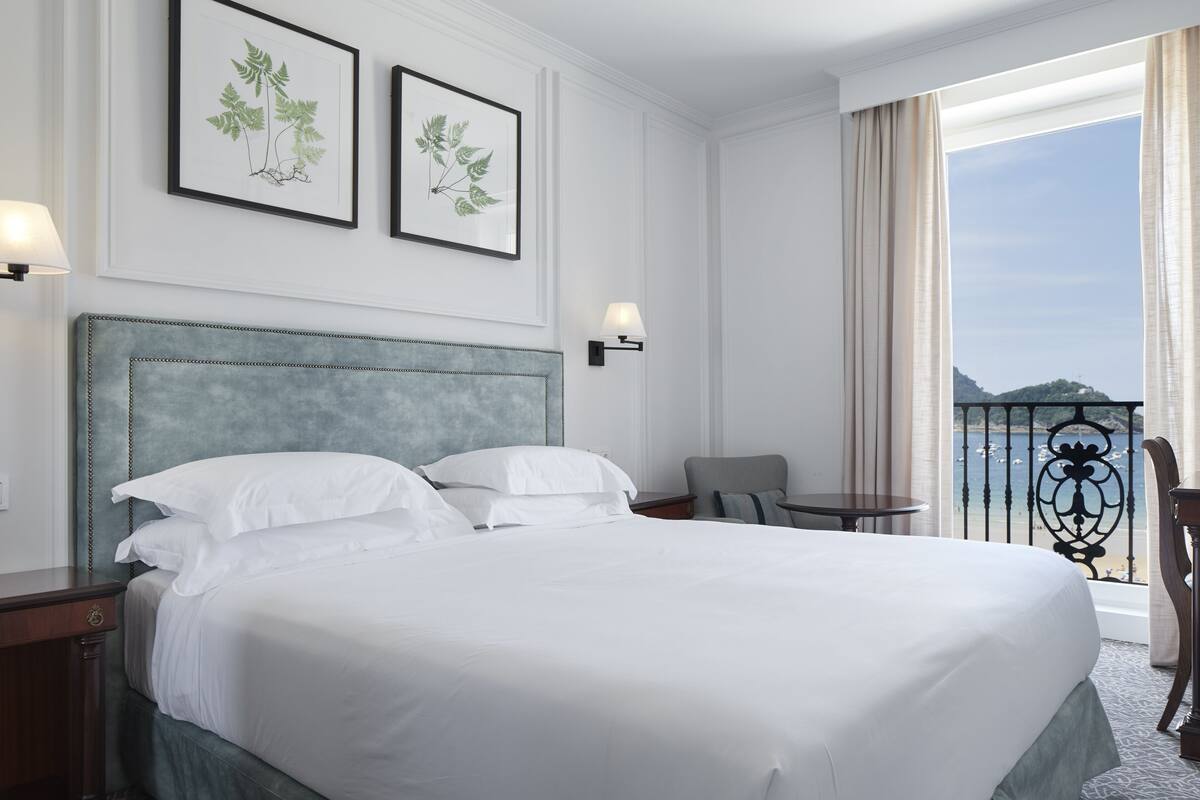 Hotelkamer met een groot bed, twee ingelijste botanische prints aan de muur, een balkon met uitzicht op zee en een kleine tafel met een stoel.