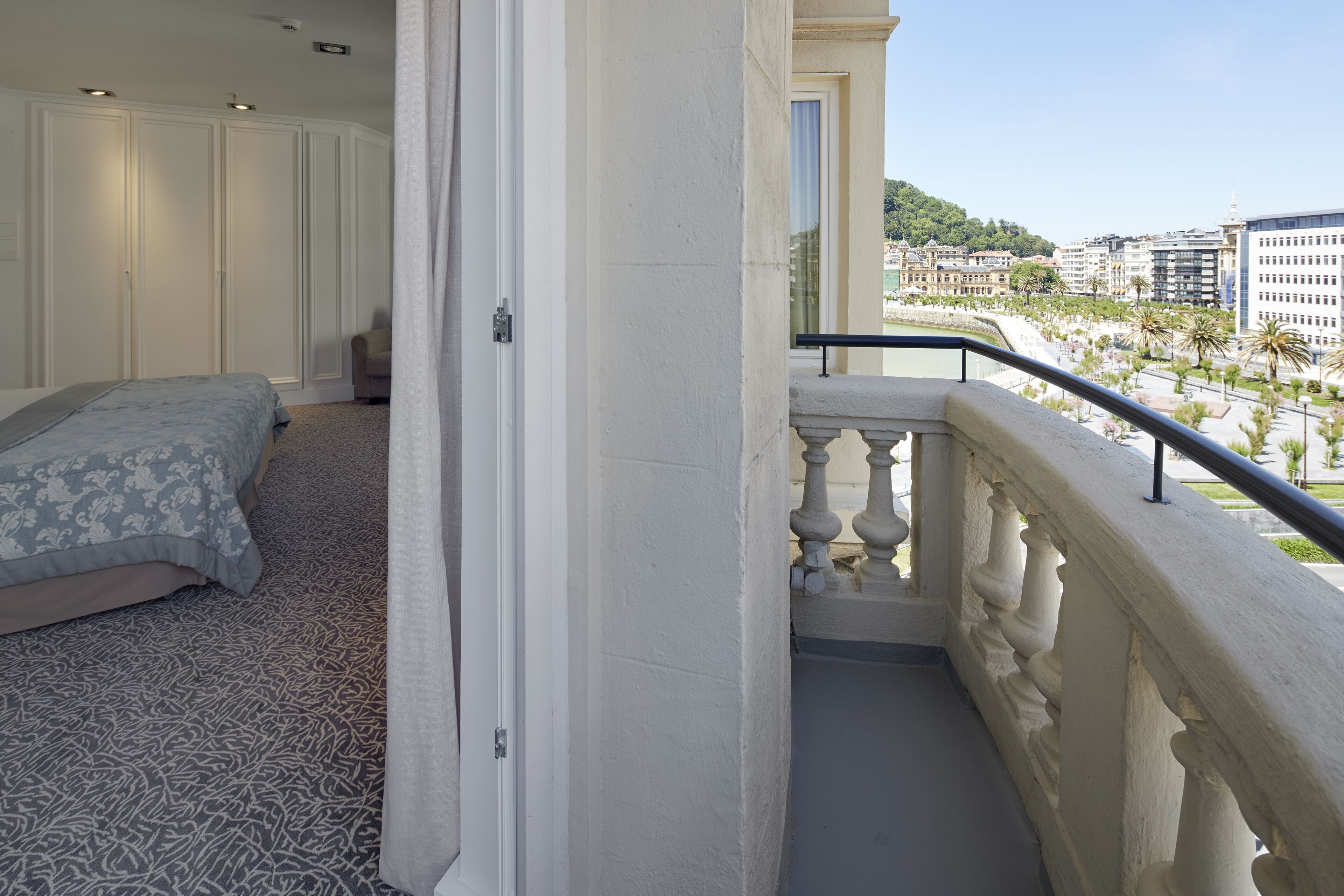 A balcony with a railing, a bed with a patterned bedspread, and a view of a cityscape with buildings and palm trees.