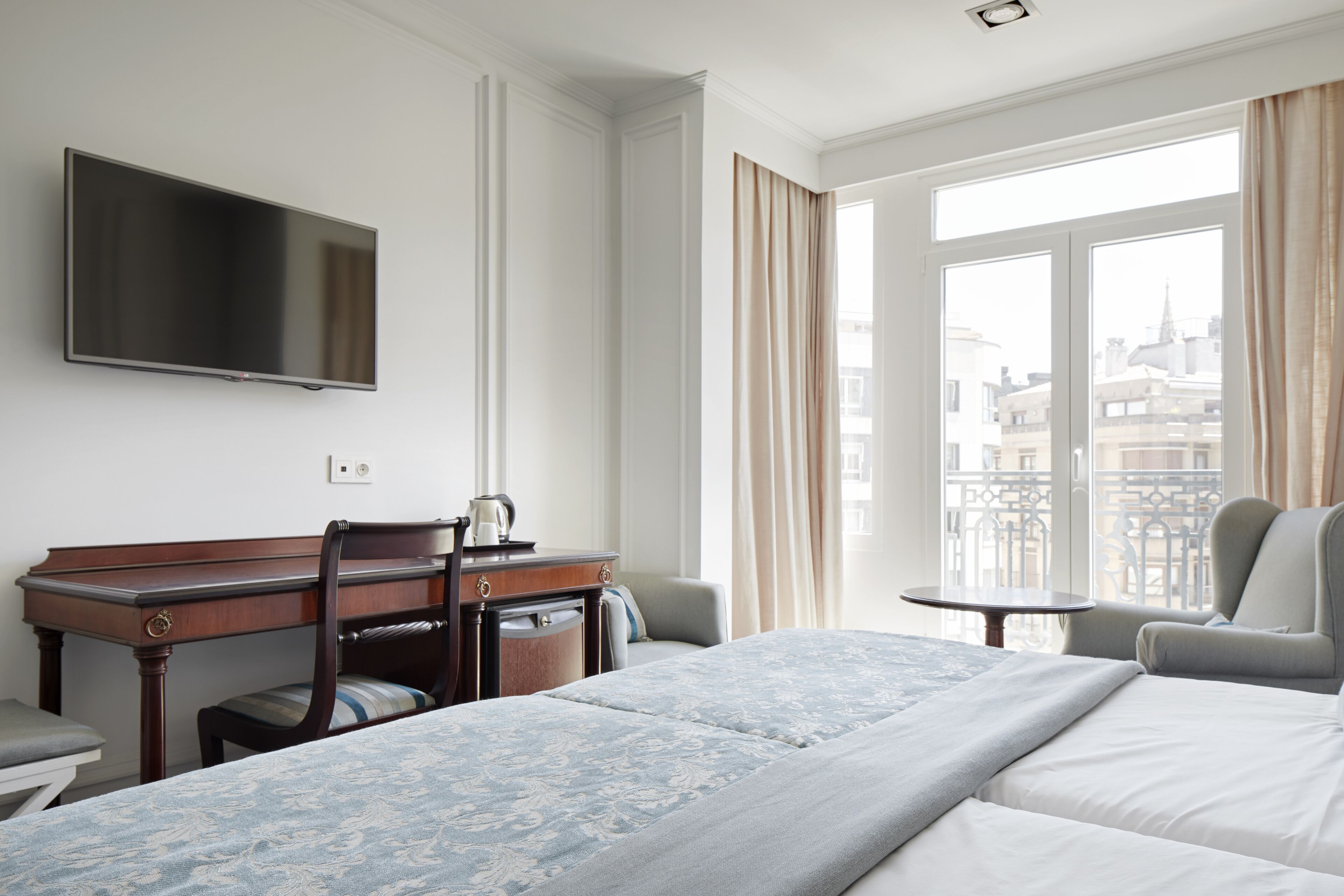 A hotel room with a bed, a desk with a chair, a television, and a balcony with a view of the city.
