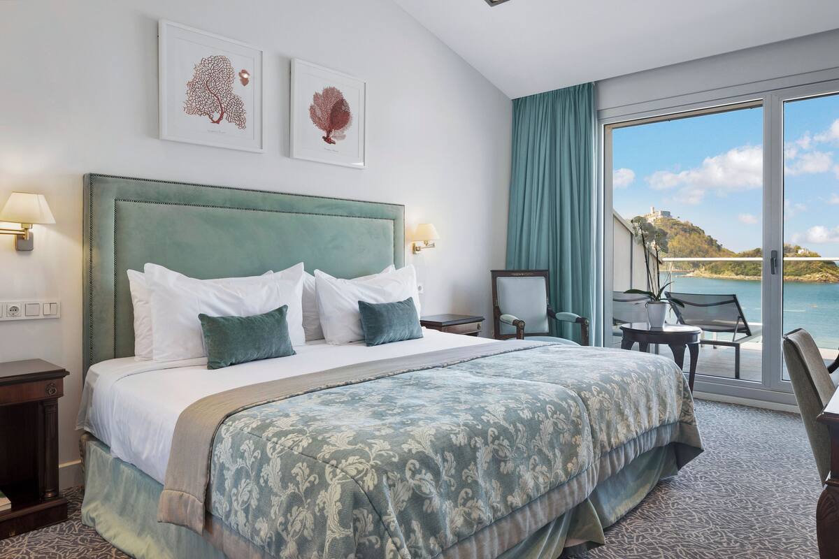 Een hotelkamer met een groot bed, twee wandlampen, uitzicht op zee en een balkon met een tafel en stoelen.