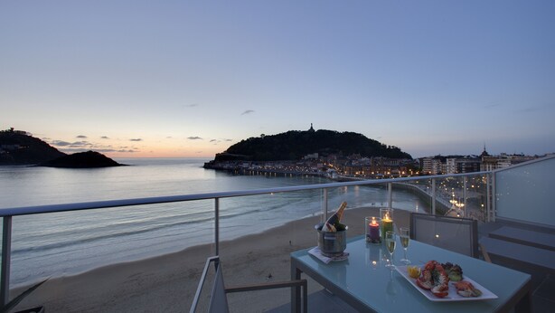 Een balkon met een eettafel, met uitzicht op een strand en een stadsbeeld in de schemering.