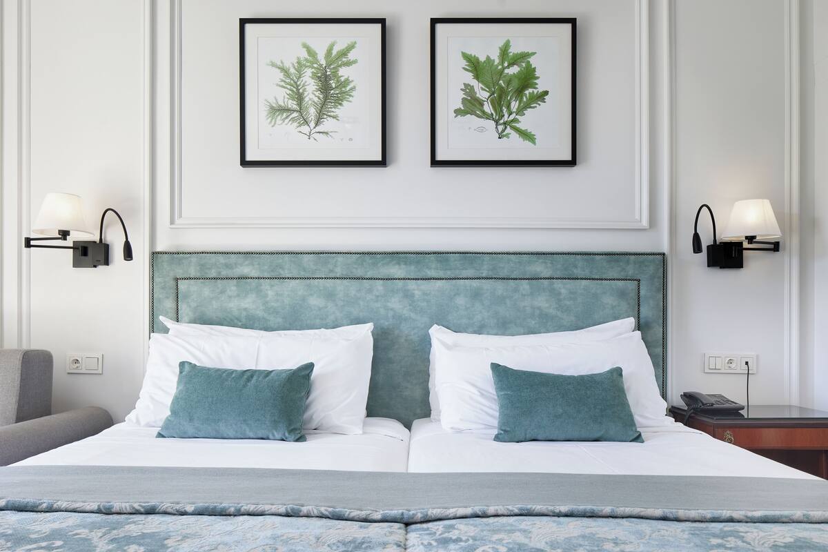 Een net opgemaakt tweepersoonsbed met witte en blauwgroene kussens, een blauwgroen hoofdeinde, en twee ingelijste botanische prints erboven.