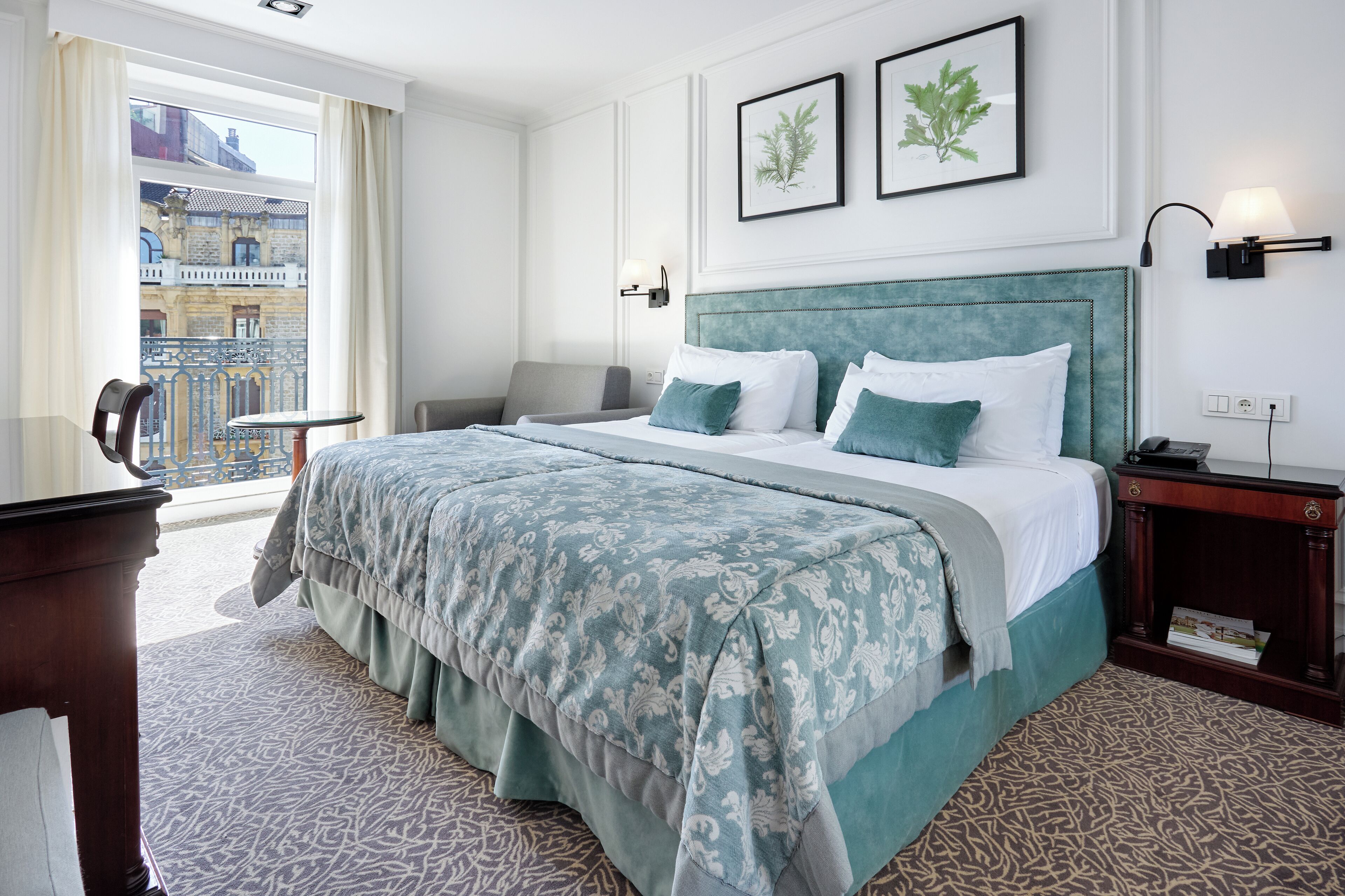 A hotel room with a large bed, a desk, a chair, a balcony with a view of buildings, and two framed pictures on the wall.