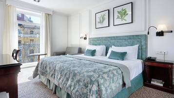 A hotel room with a large bed, a desk, a chair, a balcony with a view of buildings, and two framed pictures on the wall.