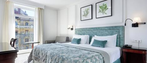 A hotel room with a large bed, a desk, a chair, a balcony with a view of buildings, and two framed pictures on the wall.
