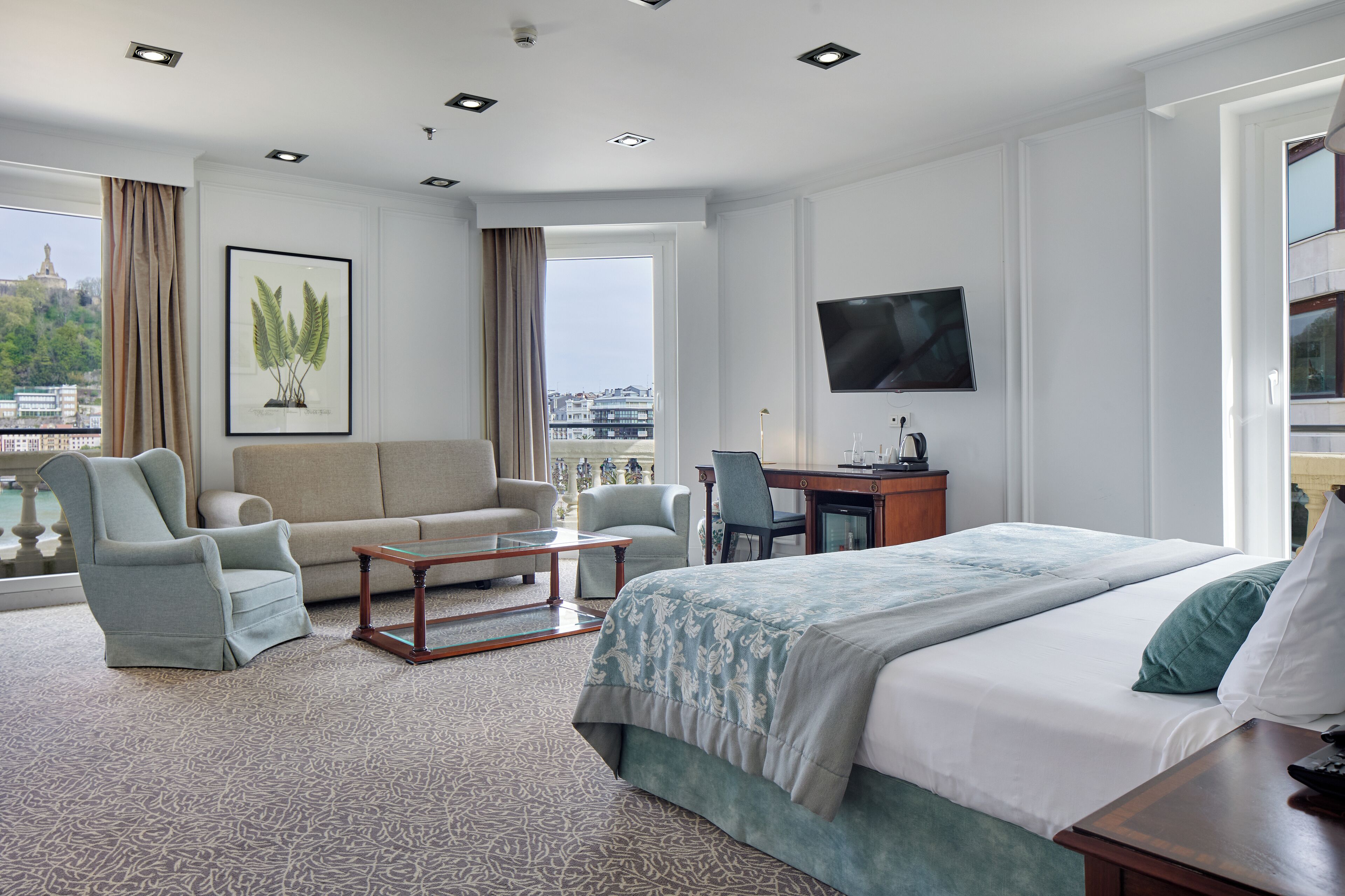 Une chambre d’hôtel comprenant un lit, deux fauteuils, un bureau, une télévision et une grande fenêtre donnant sur un paysage urbain.