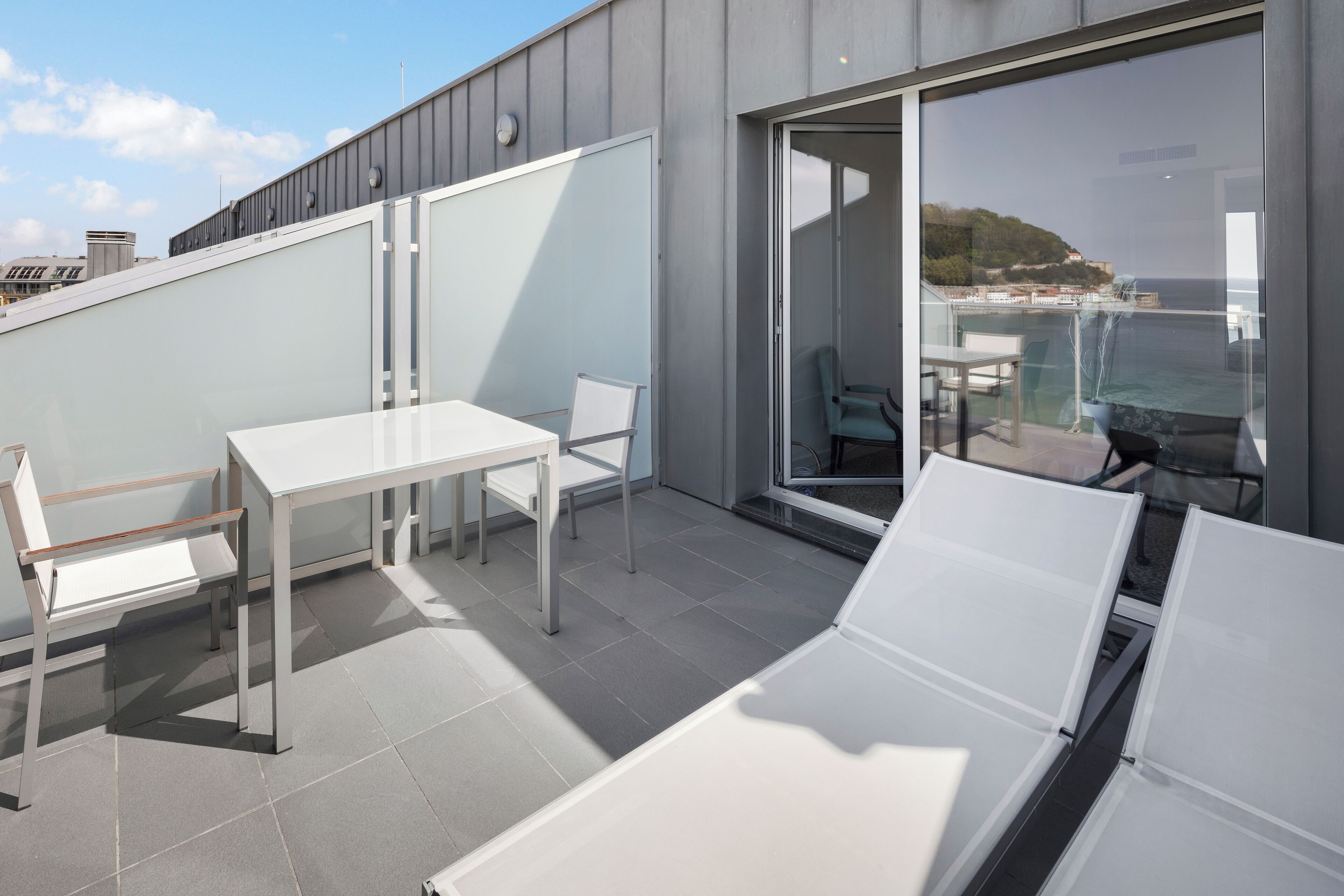 Una terraza moderna al aire libre con una mesa y sillas blancas, un ventanal de vidrio que da a un balcón con vistas al mar y una colina.