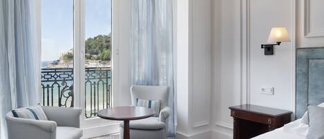 A hotel room with a bed, a chair, a small table, and a view of the sea through large windows.