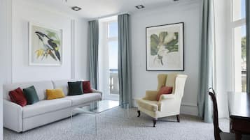 A modern living room with a white sofa, a glass coffee table, and a cream-colored armchair. There are two framed botanical prints on the walls and a large window with curtains.