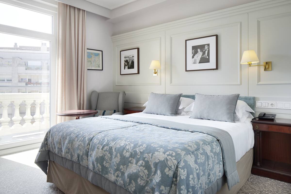 Hotelkamer met een groot bed, een bureau en een stoel. Twee ingekaderde foto’s hangen aan de muur, er staat een lamp op het bureau en er is een raam met gordijnen.