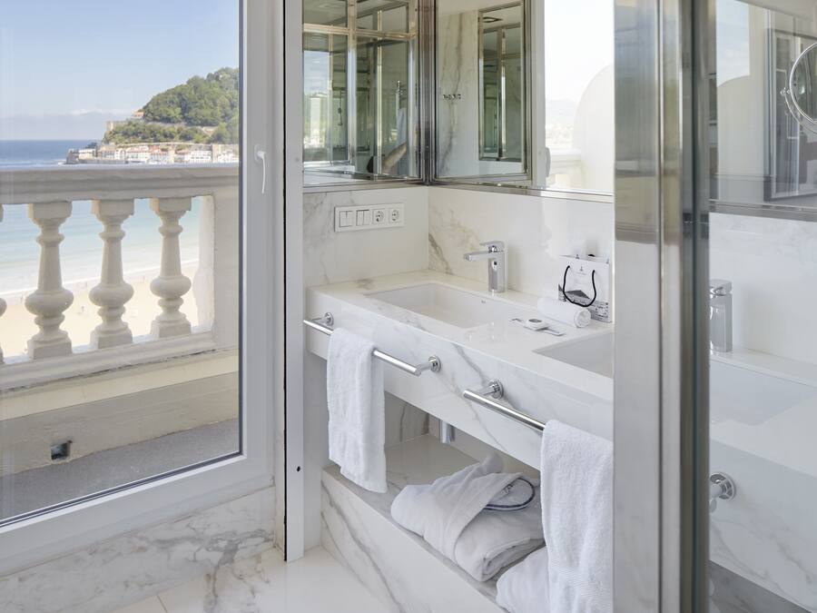 Et moderne badeværelse med et stort spejl, et badekar og en brusekabine. Det har en balkon med udsigt til havet og en bygning i baggrunden.