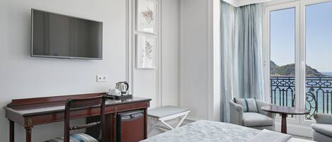 A hotel room with a bed, a desk with a chair, a small table, a TV mounted on the wall, and a balcony with a view of the sea.