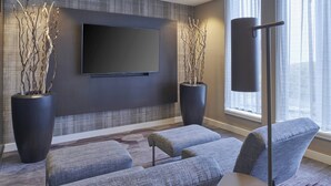 A modern living room with a flat-screen TV, two large potted plants, a floor lamp, and grey upholstered sofas. - Courtyard by Marriott Detroit Troy (Troy)