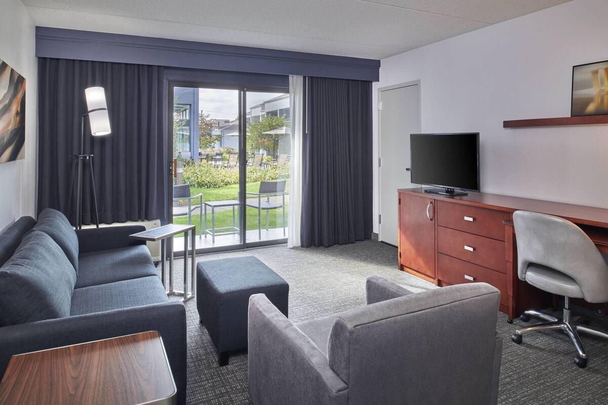 A modern hotel room with a gray sofa, a wooden desk, a television, and a view of the outdoor garden.
