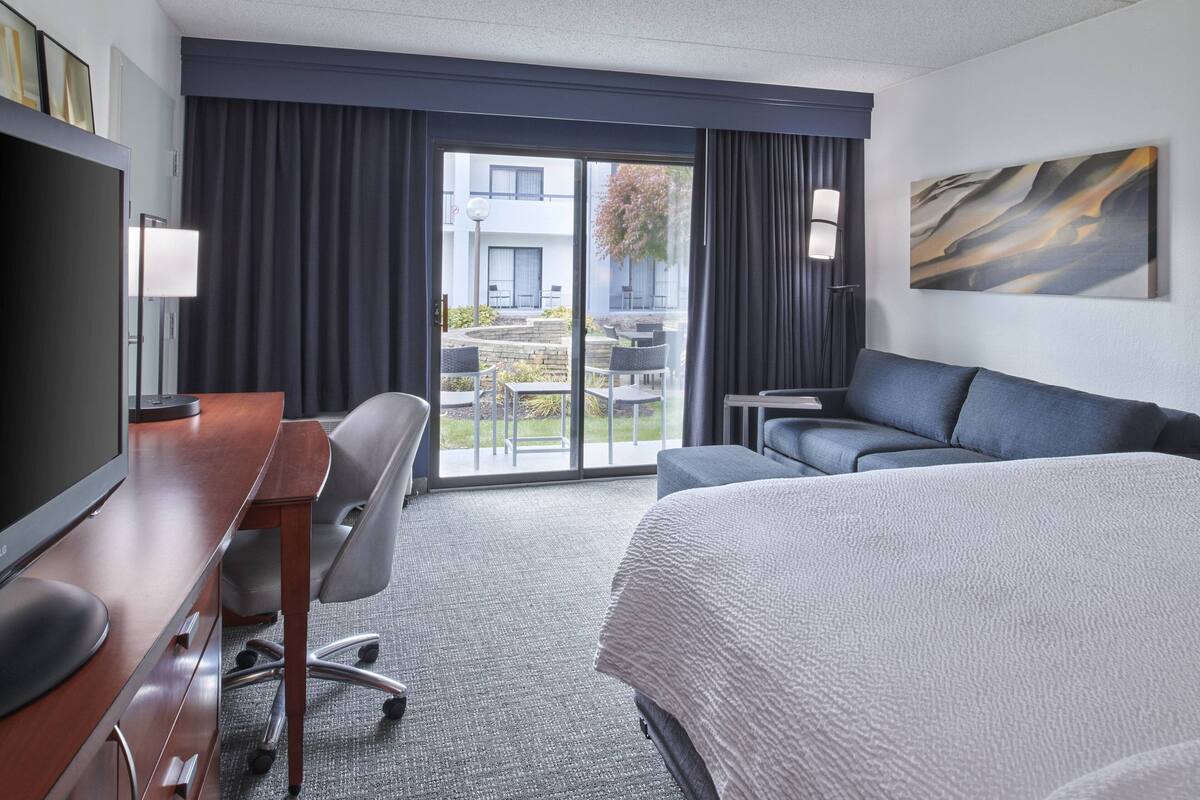 A hotel room with a large bed, a desk with a computer, a chair, a sofa, and a view of the outdoor area through a sliding door.