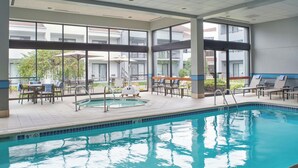 An indoor swimming pool with lounge chairs and tables, large windows, and a view of the surrounding buildings. - Courtyard by Marriott Detroit Troy (Troy)