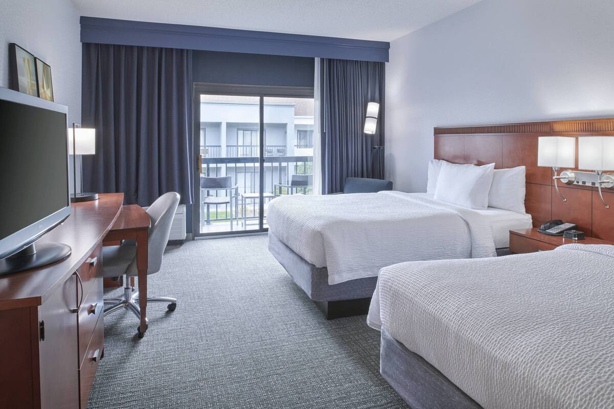 A hotel room with two beds, a desk, a chair, a TV, and a balcony with a view of the building.
