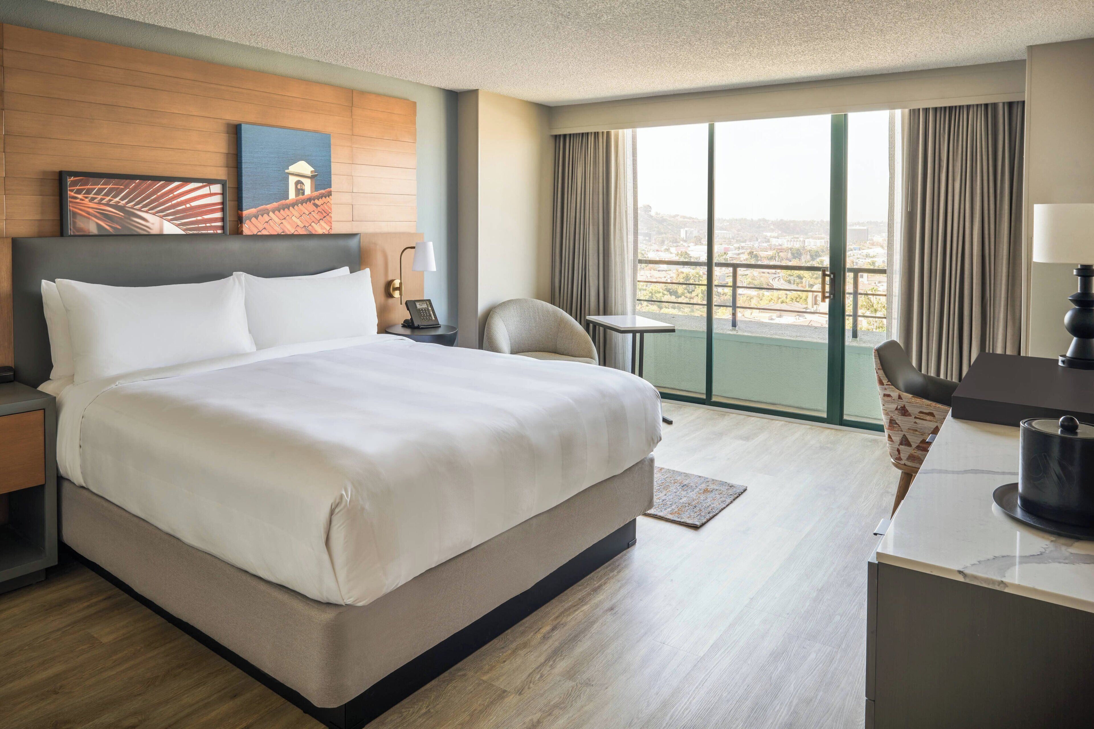 A hotel room with a large bed, a desk, a chair, a view of the city, and a balcony.