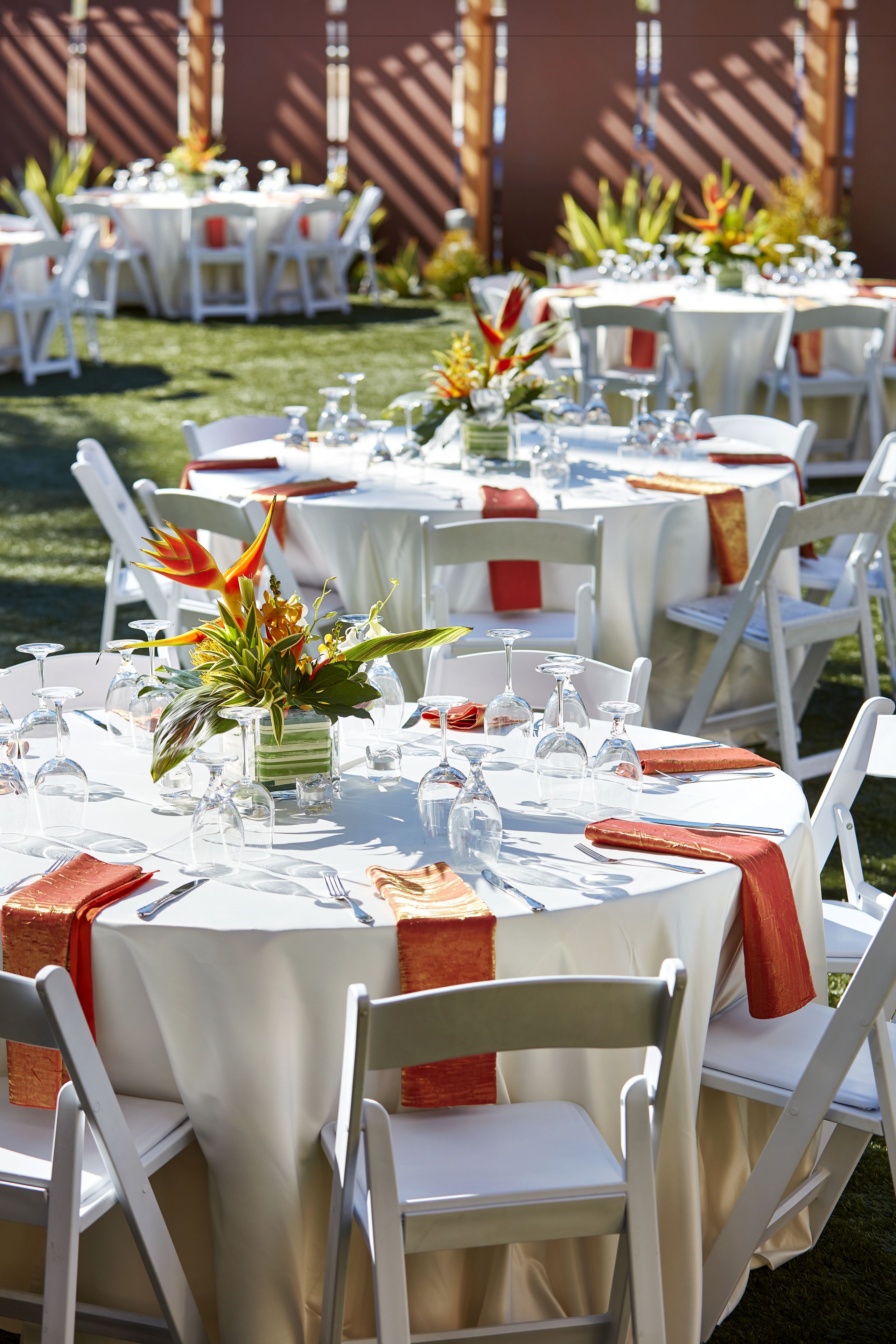 Een reeks ronde tafels buiten opgesteld, met witte stoelen en rode servetten. Elk tafel is versierd met een klein vaasje bloemen en er staat servies en bestek klaar voor een maaltijd.