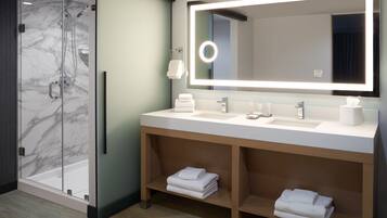 A modern bathroom with a glass shower enclosure, a large mirror with integrated lighting, a sink with a towel rack, and a vanity with shelves for towels.