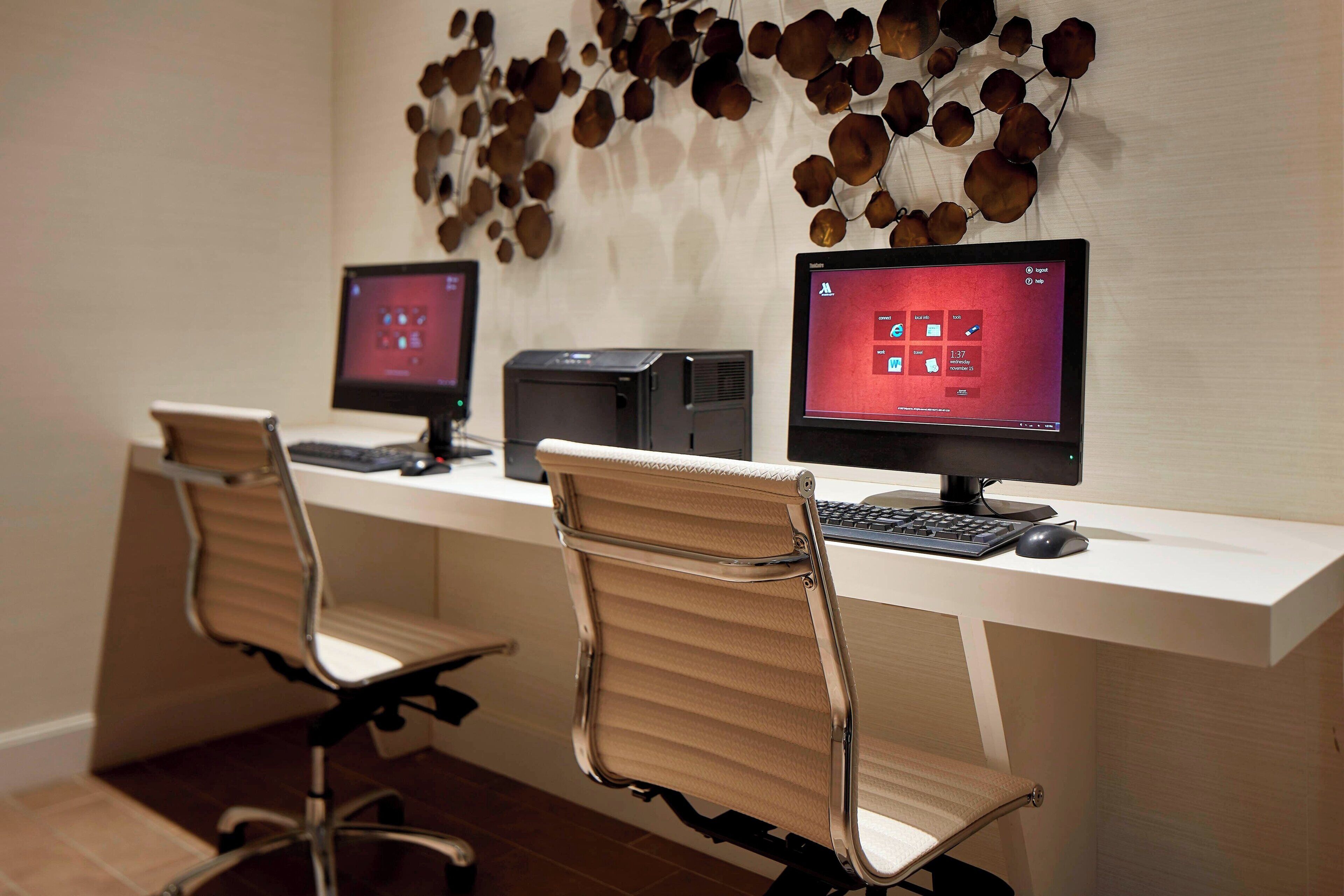 A modern office space with two desks, each with a computer monitor, keyboard, and mouse. There are two office chairs with metal frames and cushioned seats. The wall behind the desks features a decorative arrangement of brown, leaf-like shapes.
