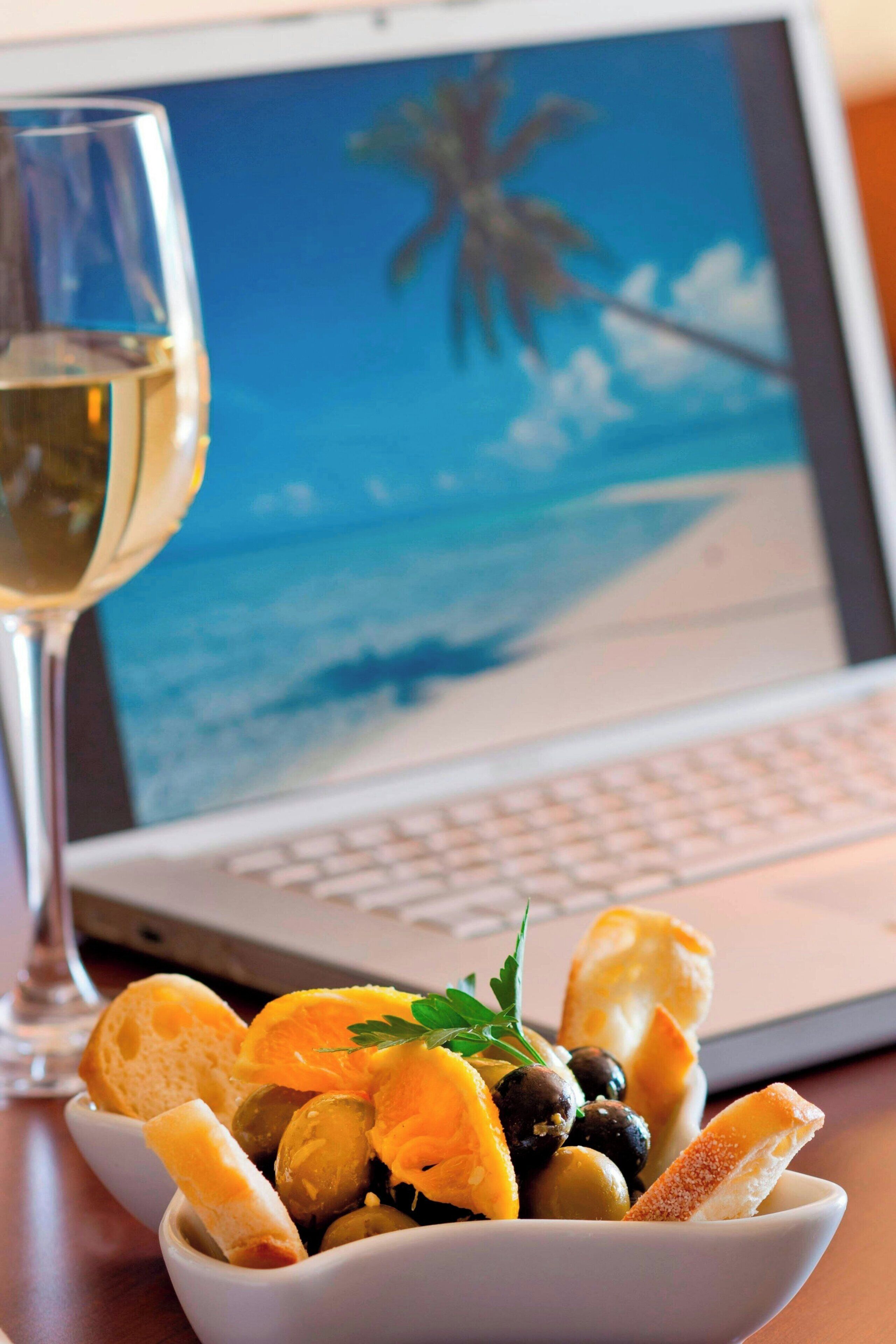 Una computadora portátil con un fondo de pantalla de playa, una copa de vino blanco y un cuenco con aceitunas y pan sobre una mesa.