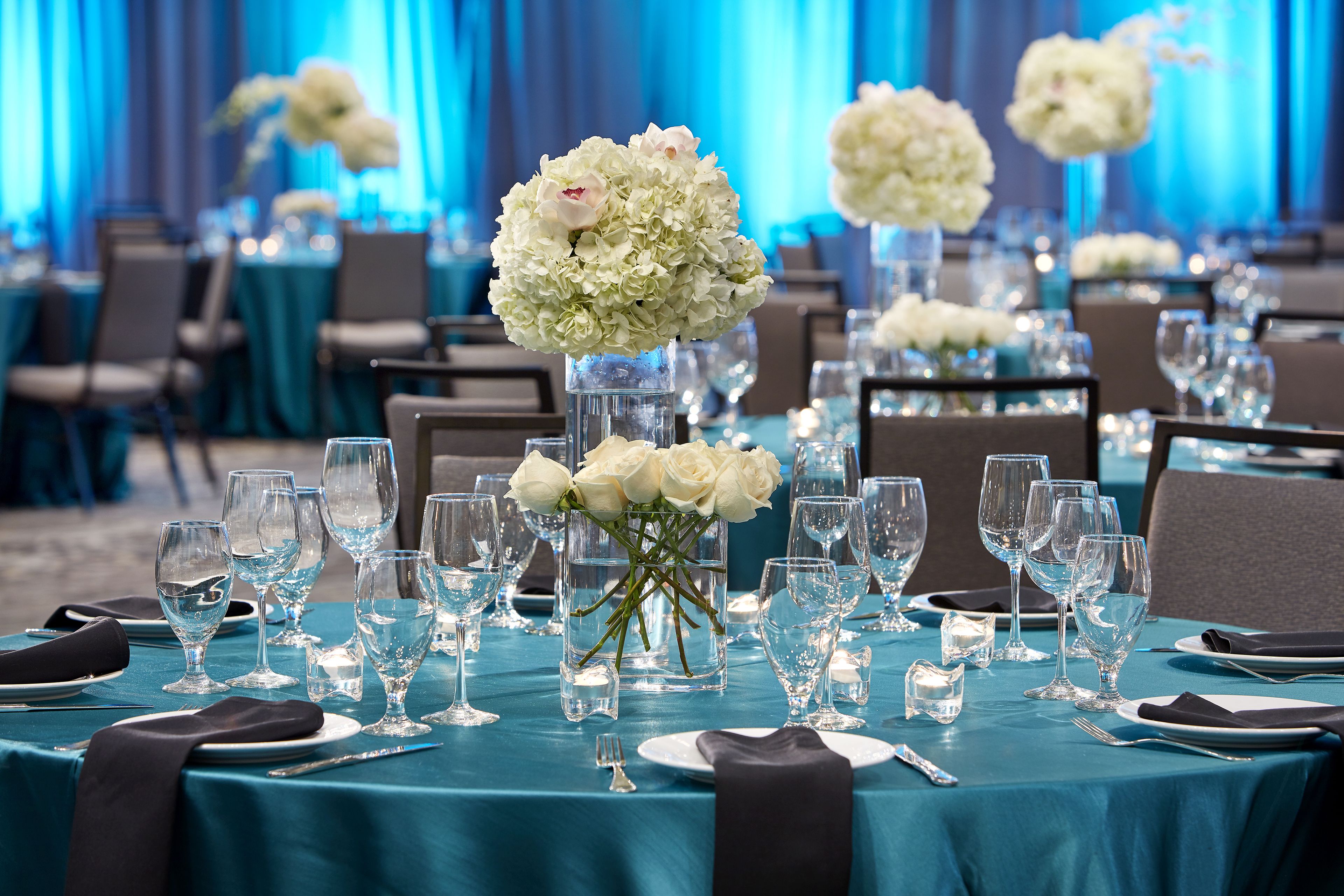 A banquet hall with tables set for a formal event, featuring clear glassware, white flowers, and dark tablecloths.
