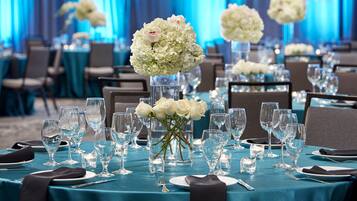 A banquet hall with tables set for a formal event, featuring clear glassware, white flowers, and dark tablecloths.