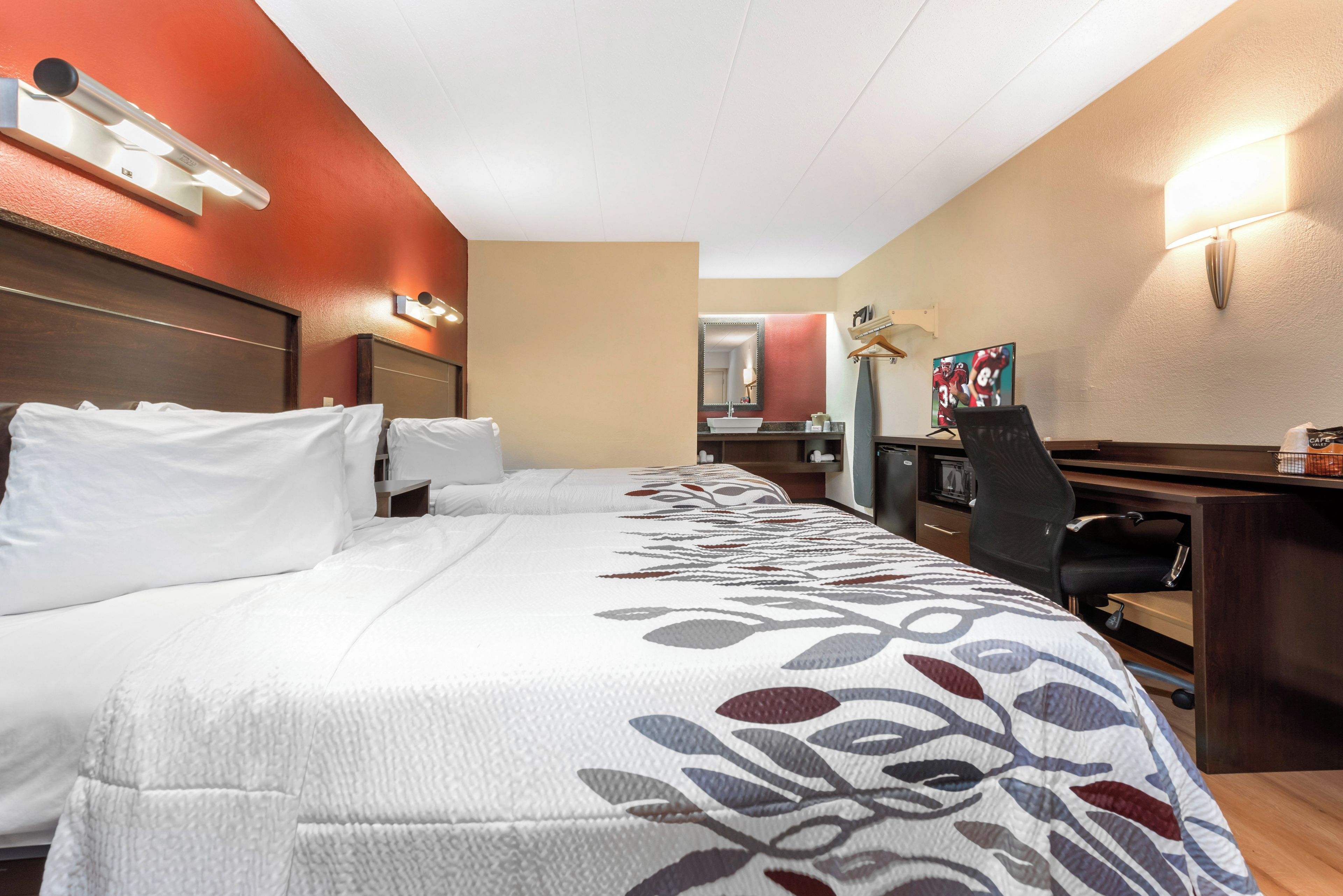 A hotel room with two beds, a desk, a chair, a mirror, and a television.