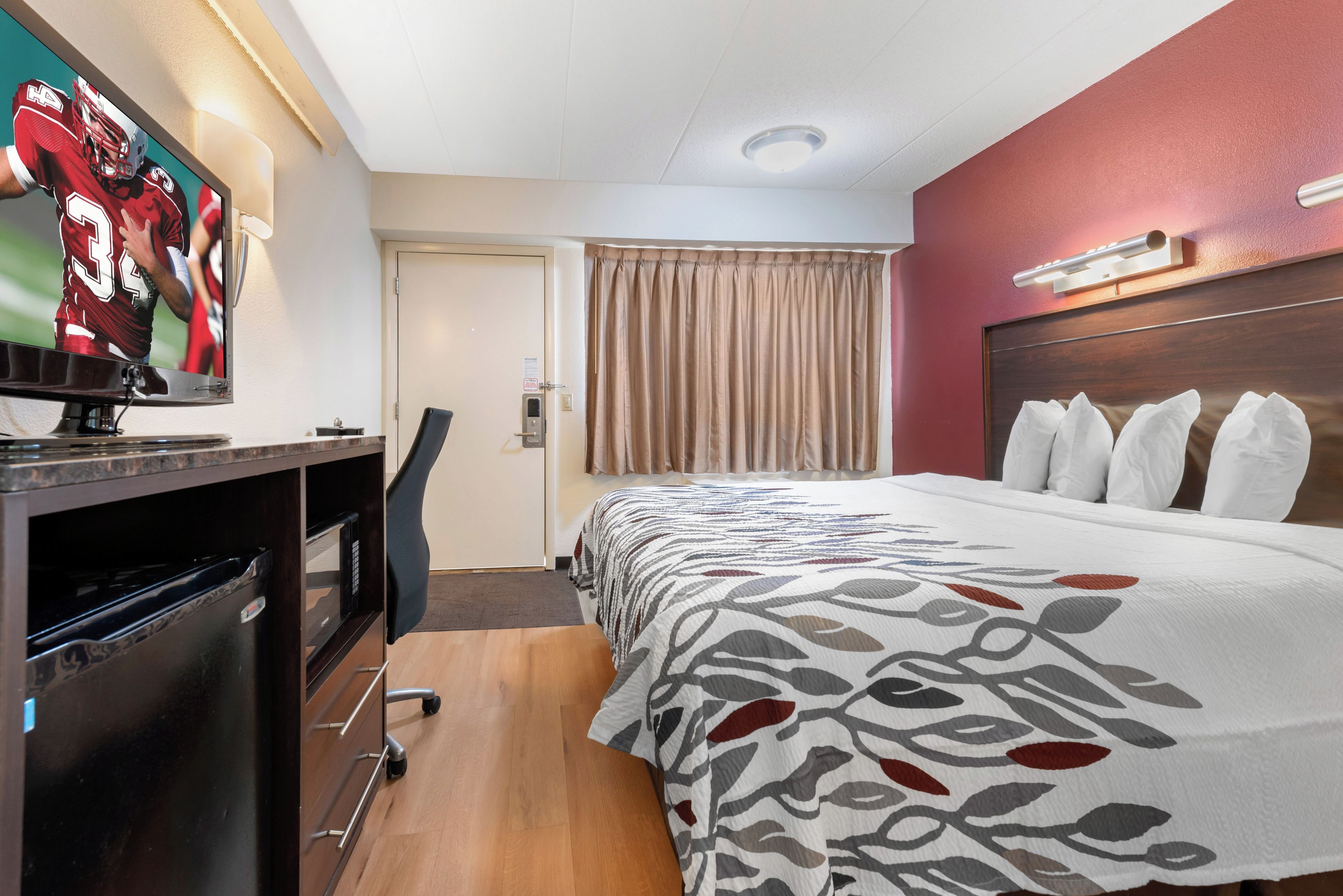 Habitación de hotel con cama, escritorio con televisor, silla, puerta y ventana con cortinas.